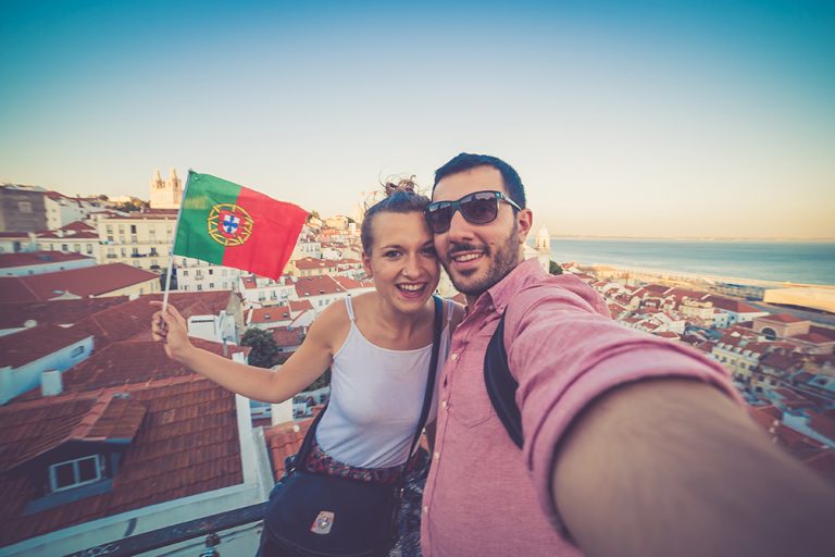 Casal de turistas tira selfie com vista para Portugal com pôr do sol ao fundo segurando bandeira